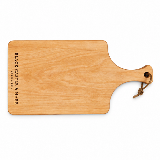 A rectangular wooden serving board with rounded corners and a handle, shown on a plain white background. The board has the text 'BLACK CASTLE & HARE FARMER'S MARKET' printed vertically on the left side and a small brown rope loop attached to the handle.