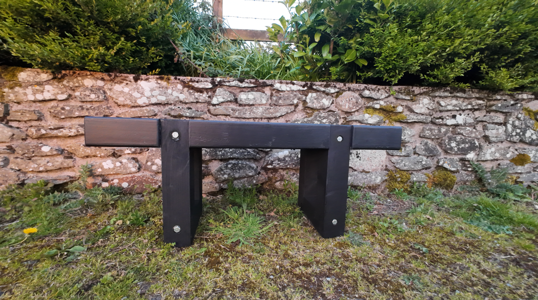 Modern two tone black wooden bench against a stone wall with greenery Side profile of the chunky wooden bench highlighting the sturdy lower stretchers and thick timber construction.