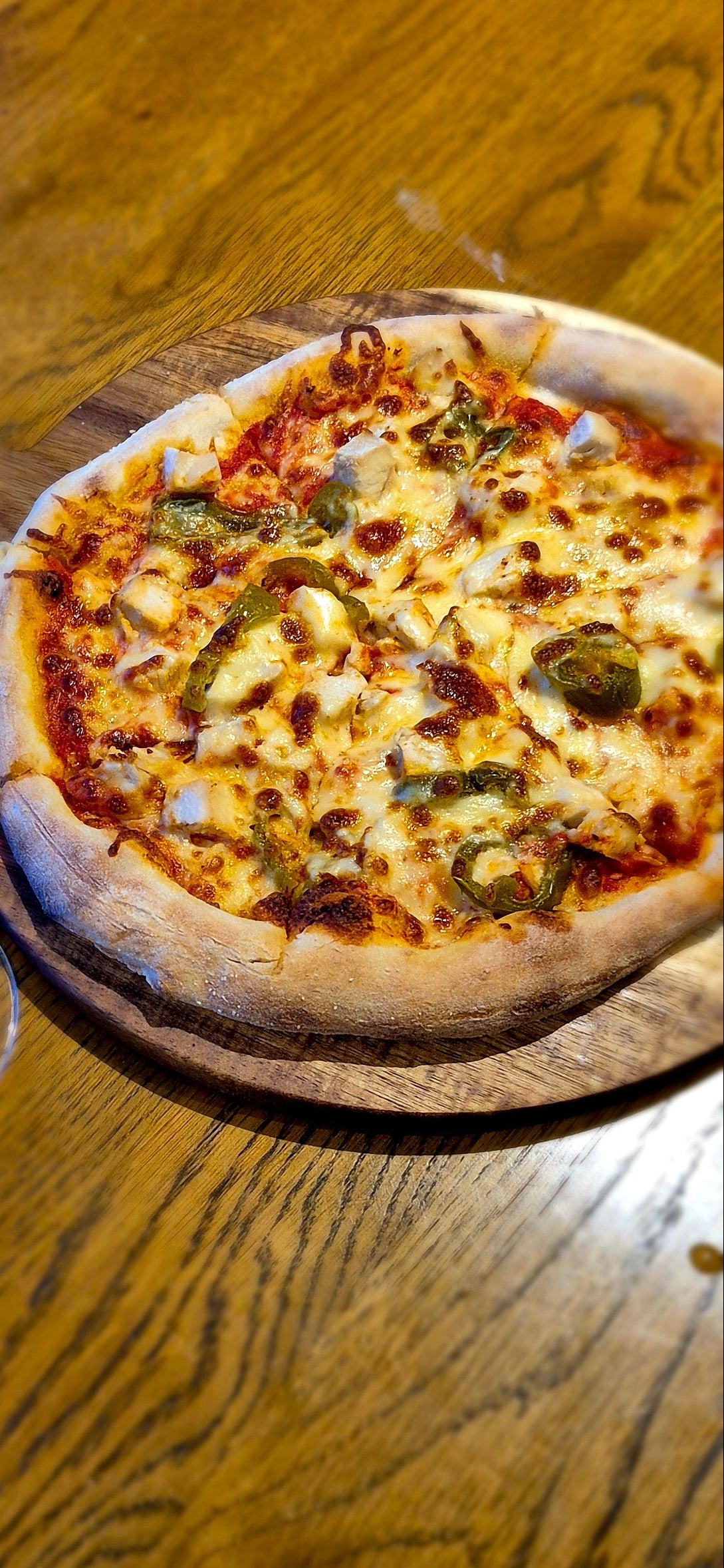 A slice of chicken and cheese pizza on a wooden board next to two glasses of red wine on a rustic wooden table.