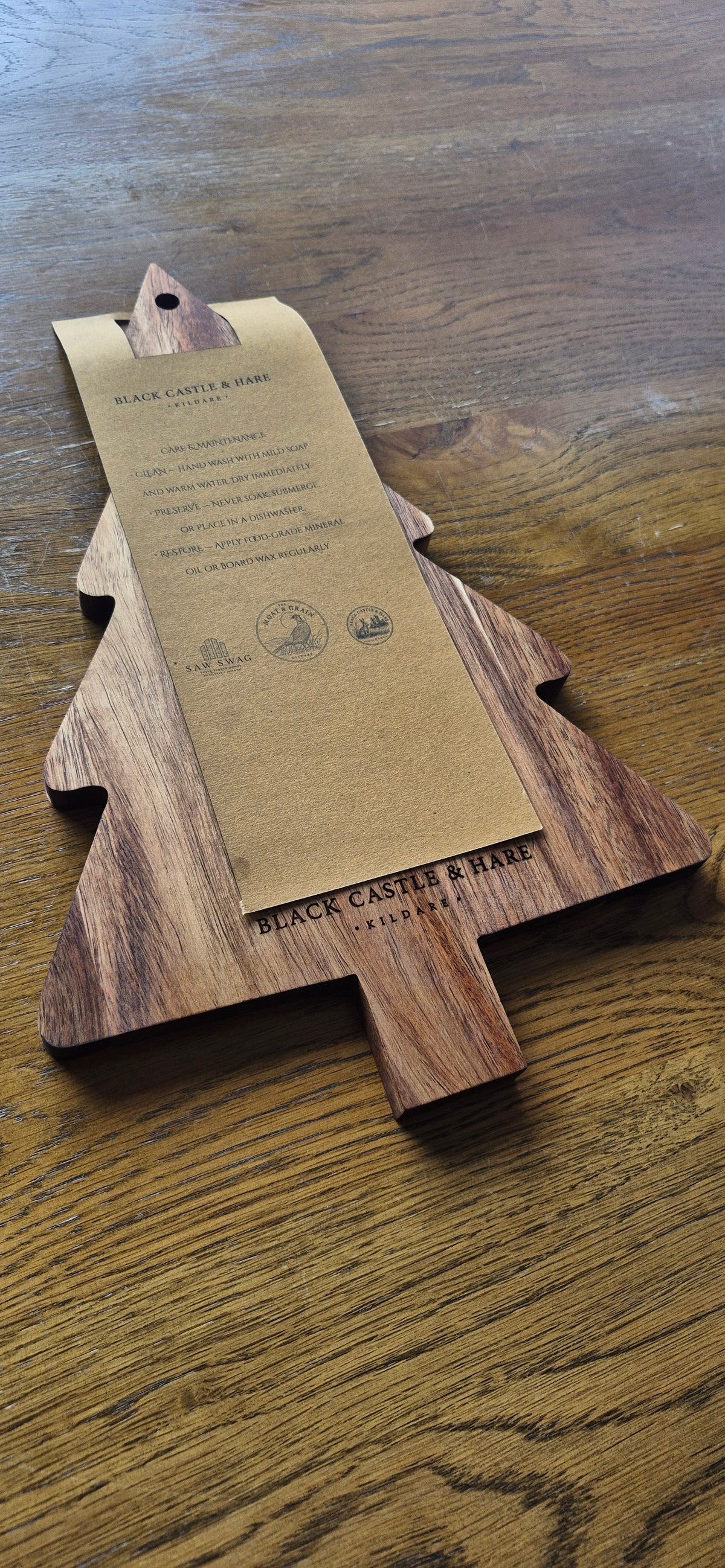 A Christmas tree-shaped wooden serving board with a brown kraft paper tag attached, placed on a wooden tabletop. The board is engraved with 'BLACK CASTLE & HARE KILDARE' and the tag shows product CARE details.