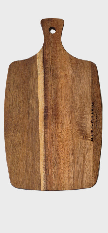 A rectangular wooden cutting board with a rounded handle and a hole at the top, featuring a natural wood grain pattern and a small brand engraving on the right side, shown against a white background.