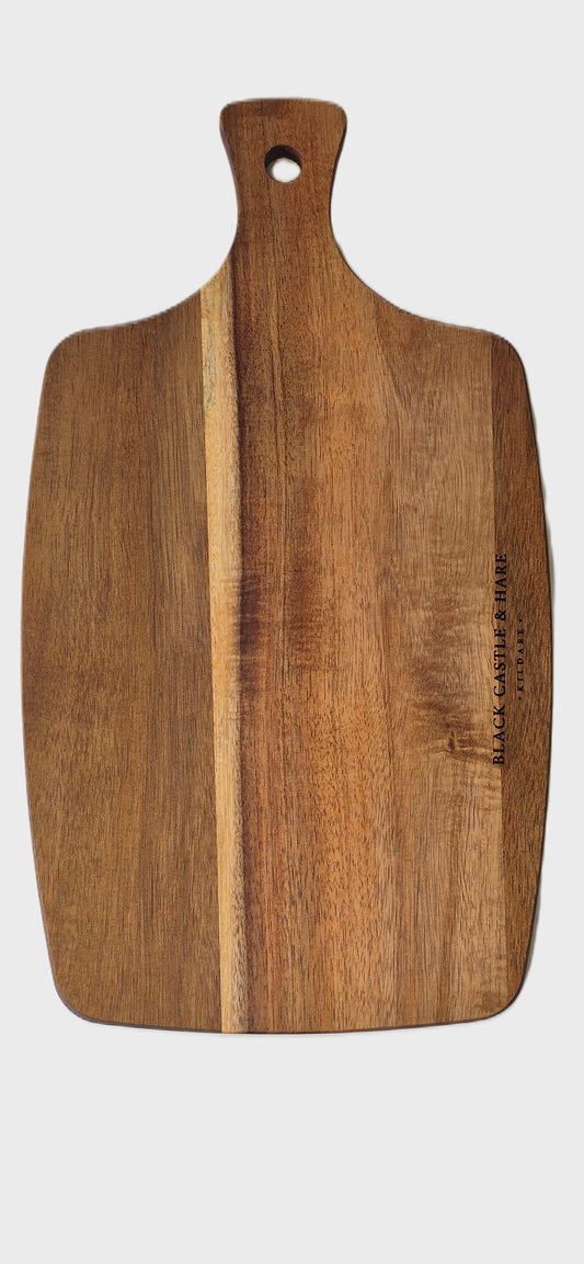 A rectangular wooden cutting board with a rounded handle and a hole at the top, featuring a natural wood grain pattern and a small brand engraving on the right side, shown against a white background.