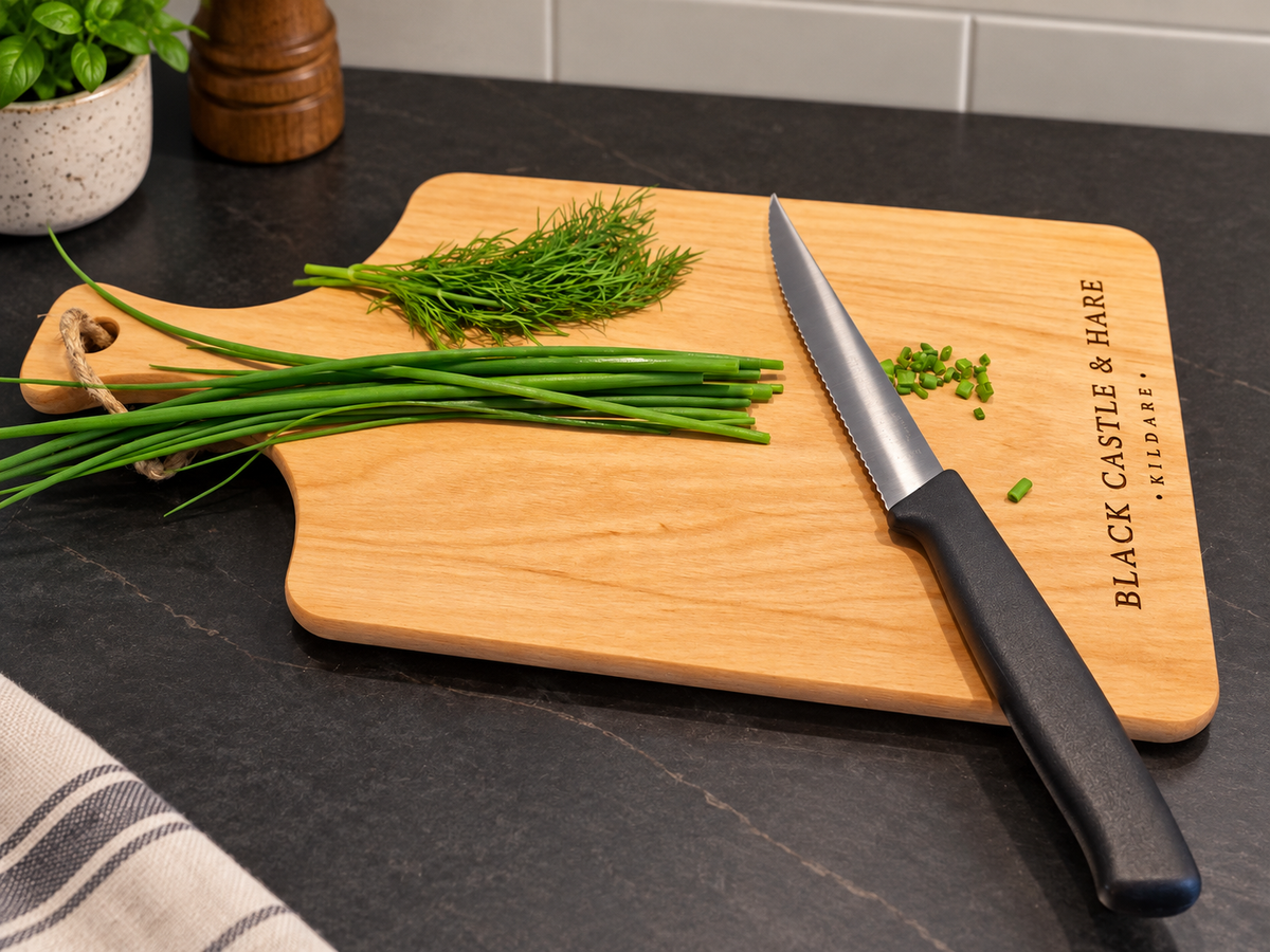 A wooden cutting board with a handle and engraved text reading 'BLACK CASTLE & HARE EST. 2024' sits on a dark countertop.