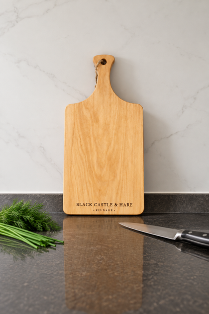 A rectangular wooden cutting board with a handle, engraved with 'BLACK CASTLE & HARE EST. 2024', standing on a dark countertop with a knife and herbs nearby against a light marble backsplash.