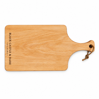 A rectangular wooden serving board with rounded corners and a handle, shown on a plain white background. The board has the text 'BLACK CASTLE & HARE FARMER'S MARKET' printed vertically on the left side and a small brown rope loop attached to the handle.