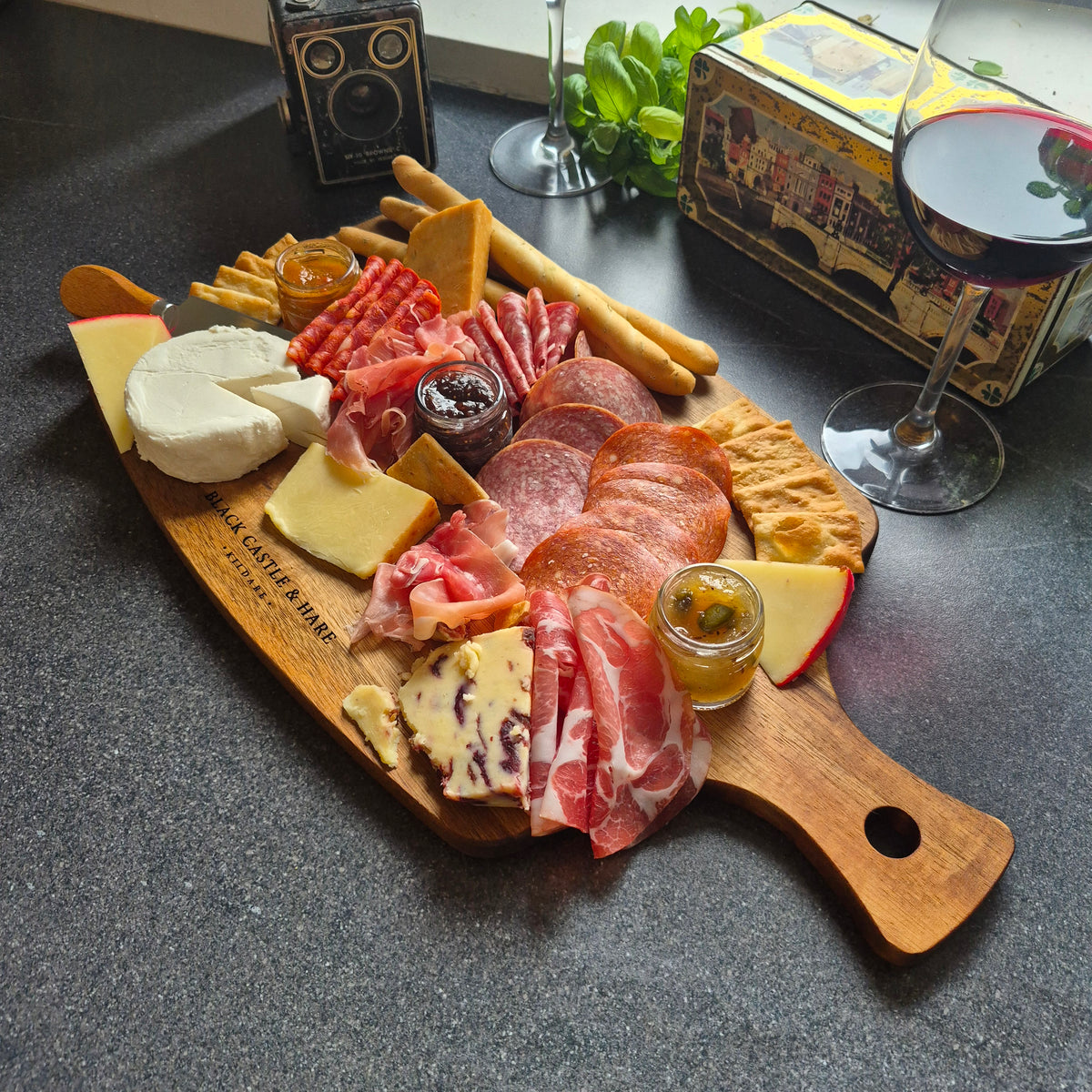 A wooden charcuterie board with an assortment of cheeses, cured meats, crackers, breadsticks, and small jars, styled on a dark countertop with a glass of red wine, a vintage camera, and a decorative box in the background aswell as the engraved branding Black Castle & Hare Kildare