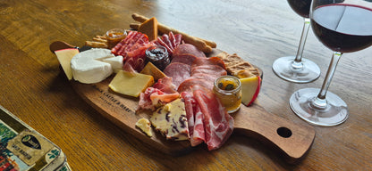 A wooden charcuterie board arranged with sliced cured meats, cheeses, crackers, and small condiment jars on a wooden table, with two stemmed wine glasses partially visible on the right. Included is the brand Black Castle & Hare Kildare.