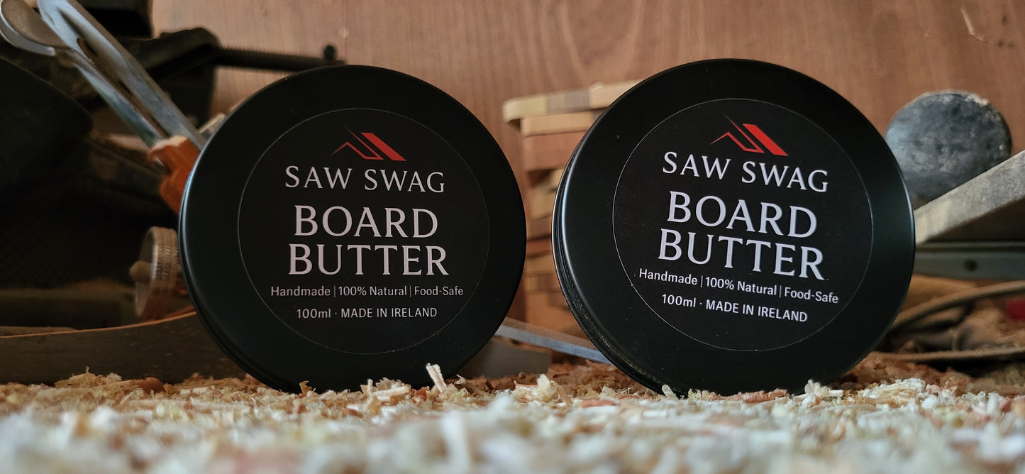 Two containers of Saw Swag Board Butter on a wooden surface with tools in the background.