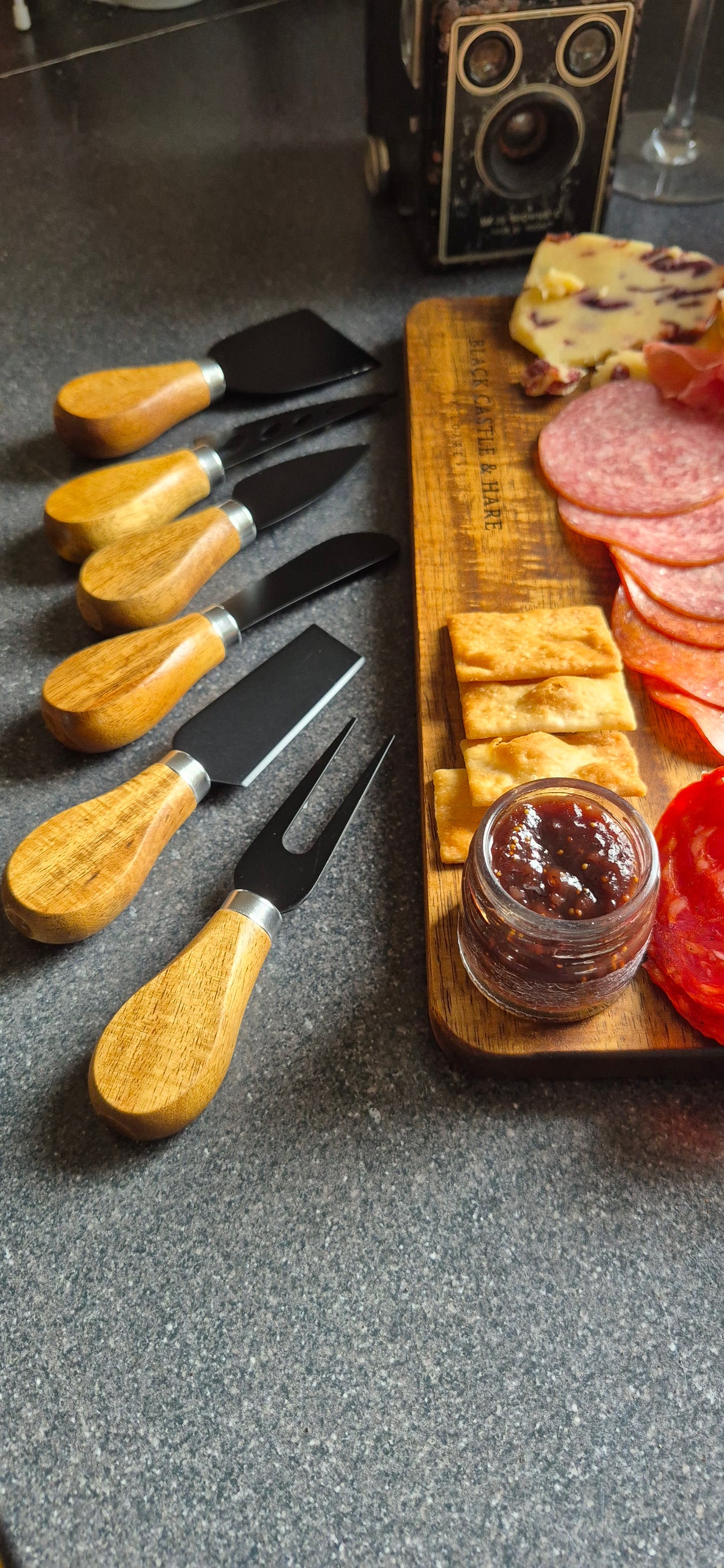 A set of cheese knives with light wood handles and black blades, arranged on a dark gray countertop next to a wooden charcuterie board with sliced meats, crackers, and a small jar of spread.