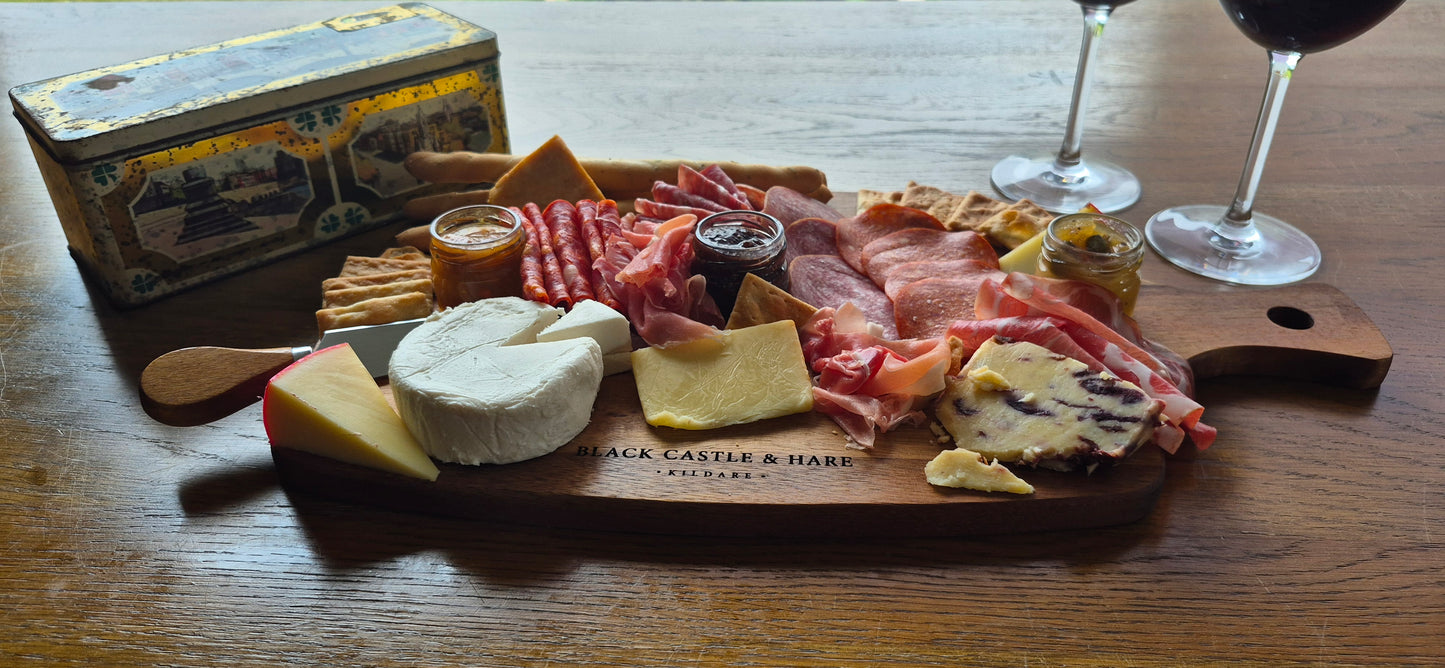 A charcuterie board spread on an oak wooden table, with the sun shining in featuring cured meats, cheeses, crackers, and small jars, with two wine glasses and an old decorative tin box nearby. The wooden serving board is branded 'Black Castle & Hare Kildare'.