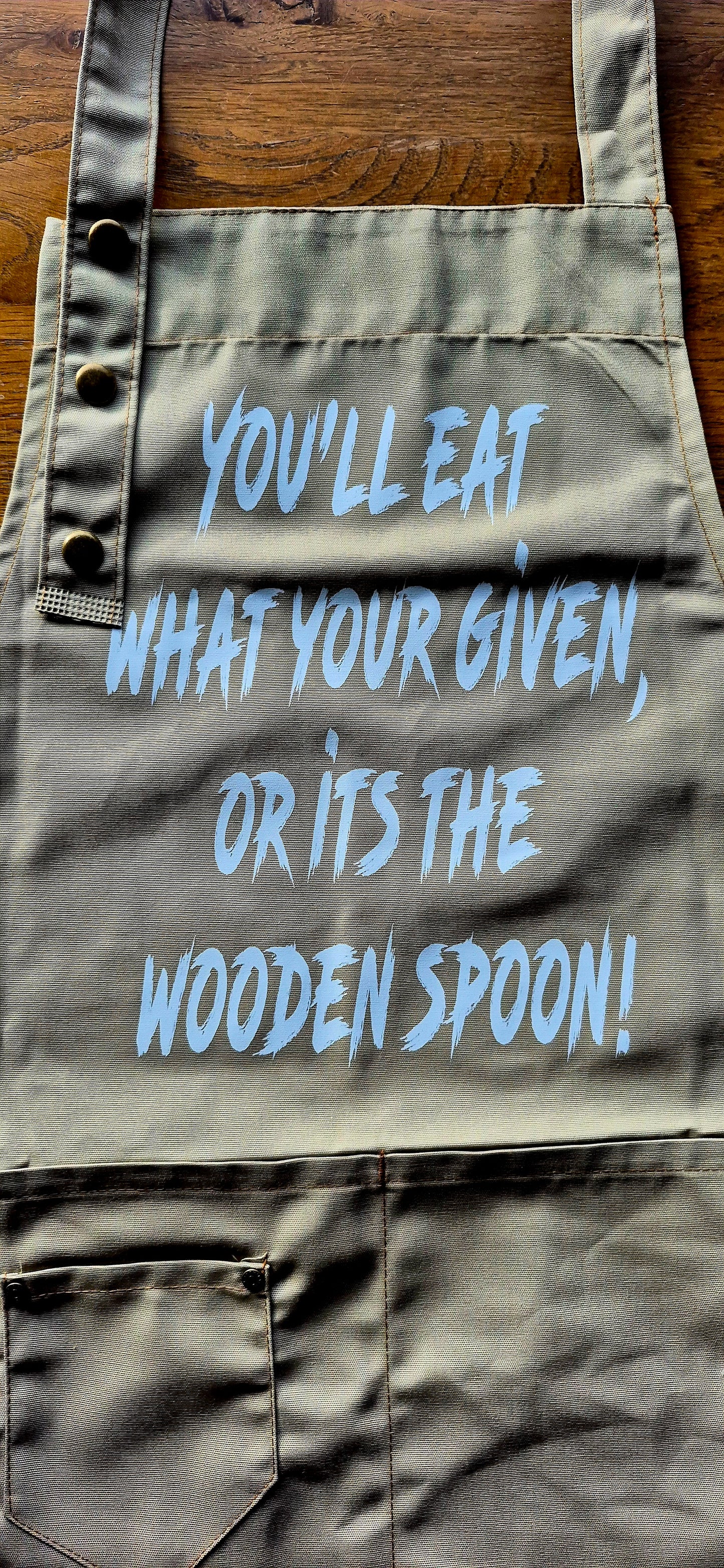 Khaki apron front view showing "Eat what you're given or it's the wooden spoon" text.