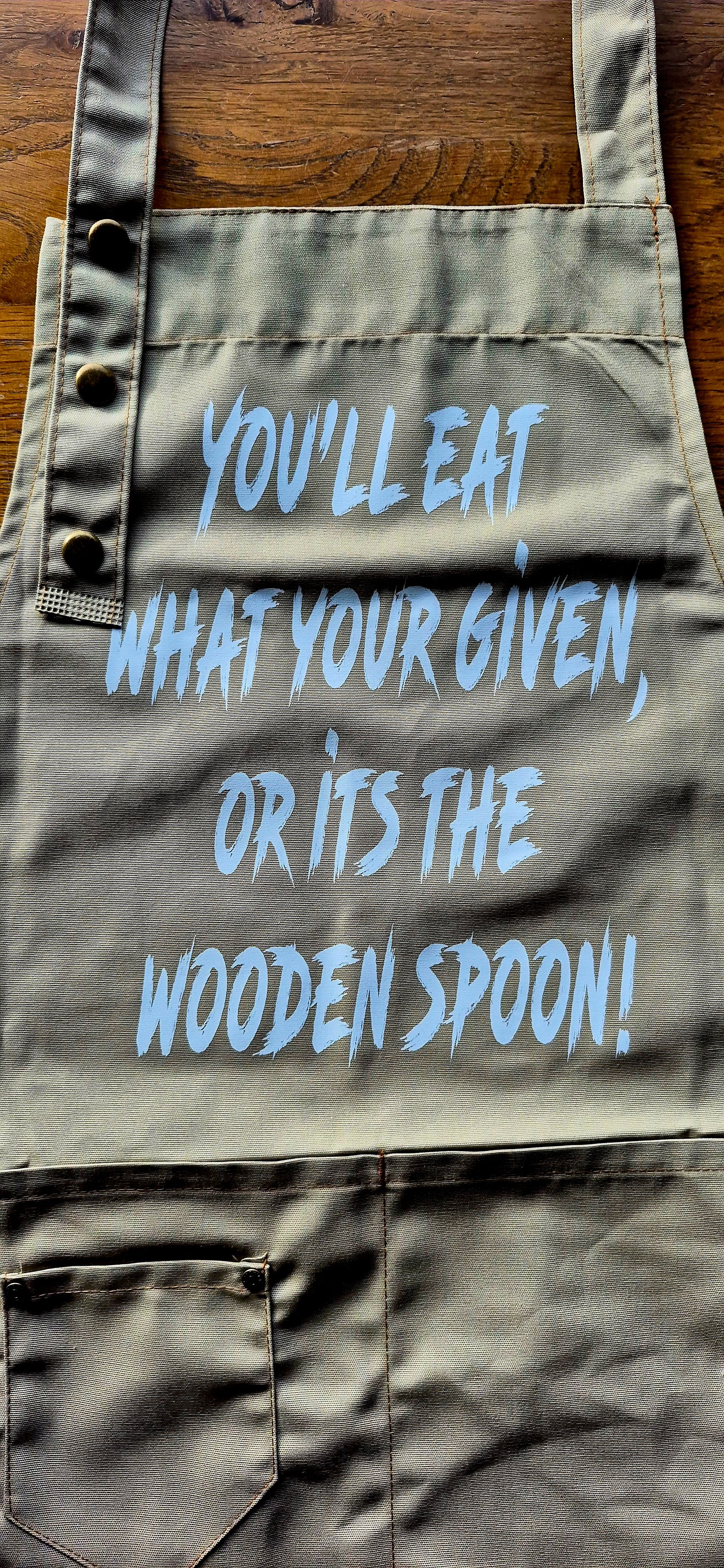 Khaki apron front view showing "Eat what you're given or it's the wooden spoon" text.