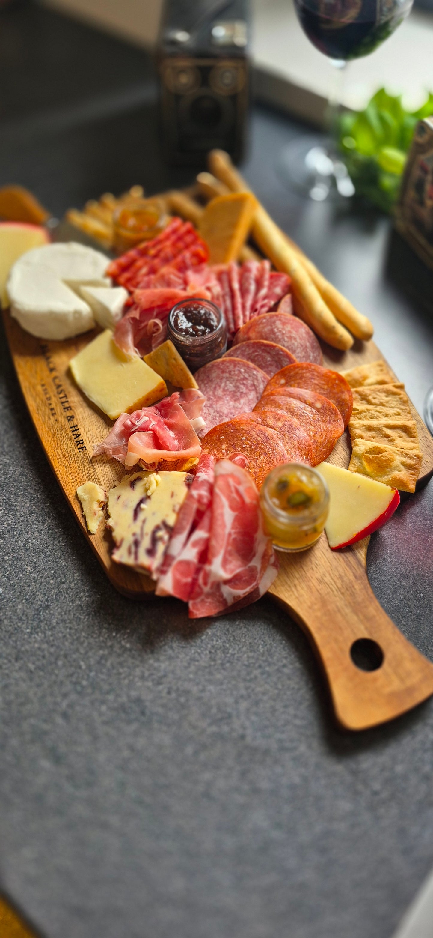 A wooden charcuterie board with the Black Castle & Hare Kildare engraved brand, filled with sliced cured meats, cheeses, crackers, and small condiment jars, photographed on a dark countertop with a softly blurred kitchen background.