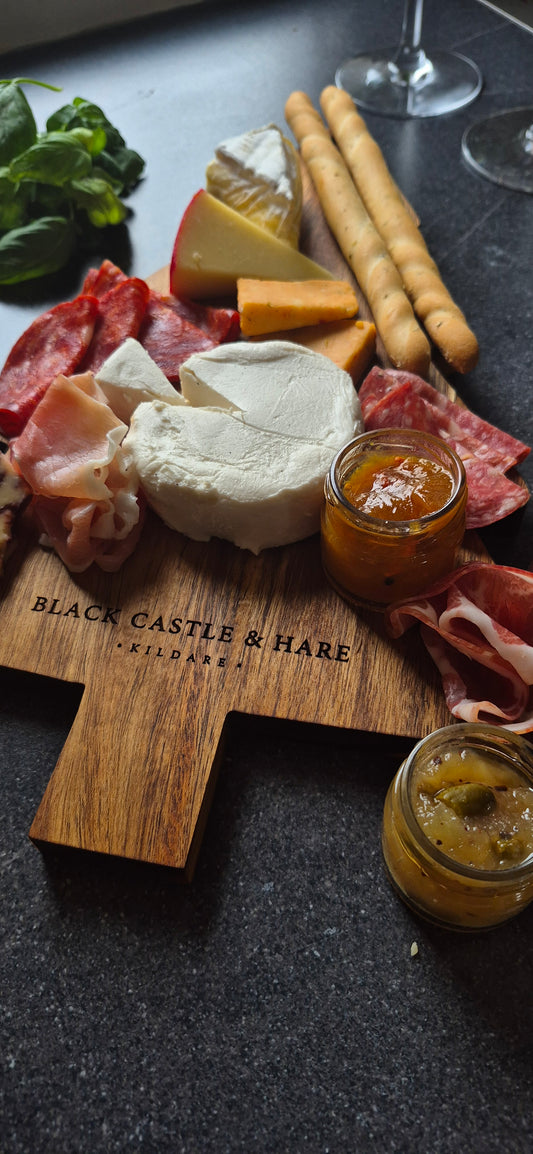A wooden charcuterie board engraved with 'BLACK CASTLE & HARE KILDARE' is arranged with sliced prosciutto, white cheese, a wedge of yellow cheese, breadsticks, and two small jars of spread on a dark tabletop.