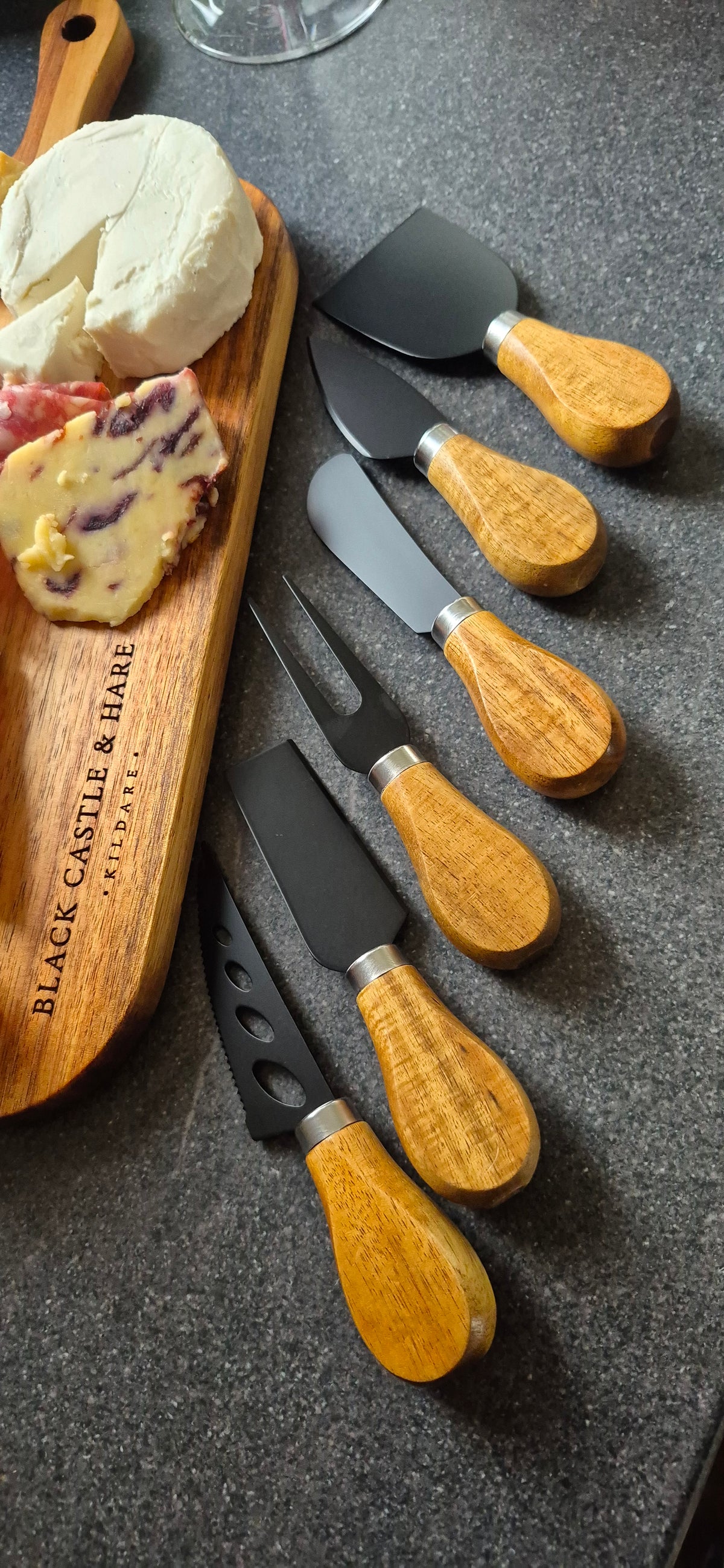 A set of cheese knives with light wood handles and black blades, arranged on a dark gray countertop next to a wooden charcuterie board with cheese.