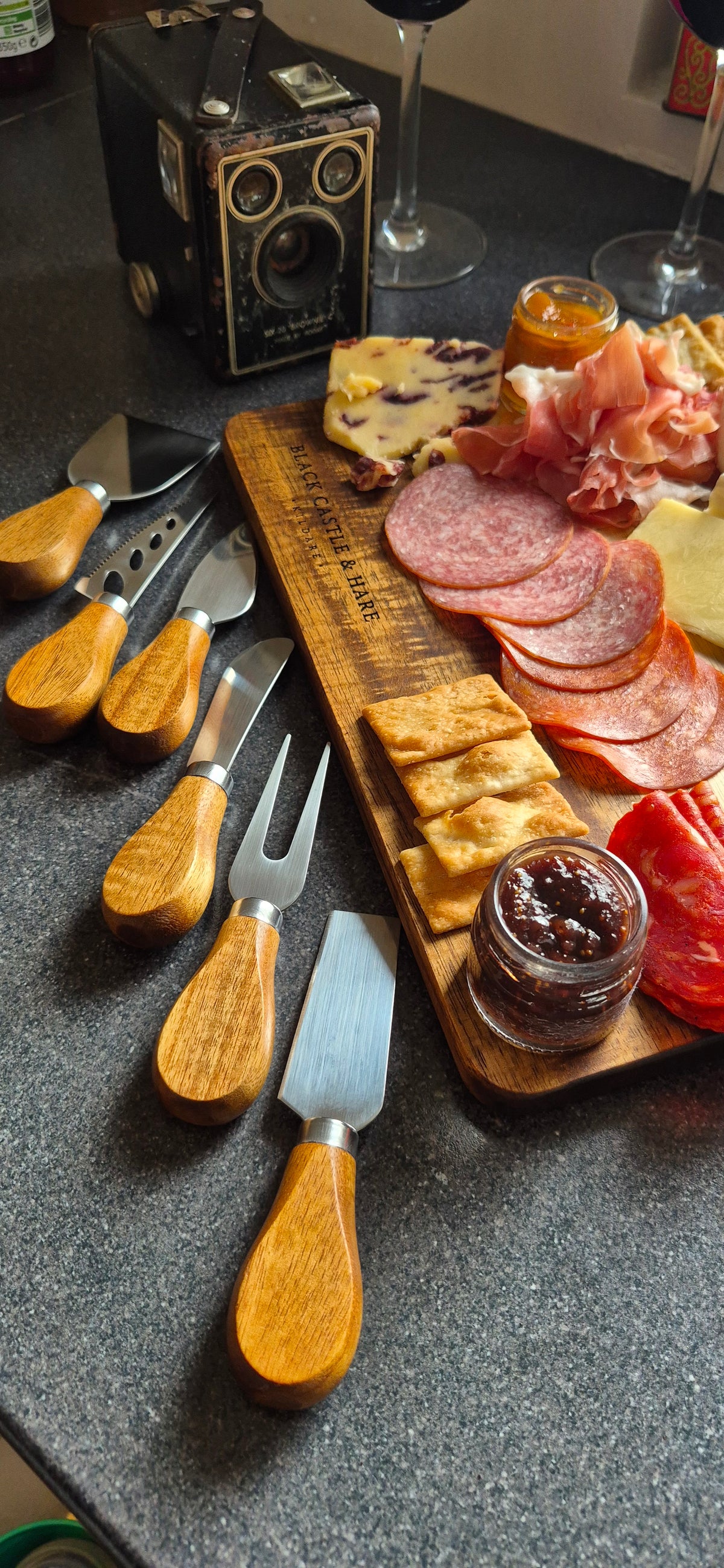 A styled charcuterie board setup on a dark countertop, featuring a wooden serving board with sliced salami, cheese, crackers, jam, and red peppers, alongside several cheese knives with light wood handles.