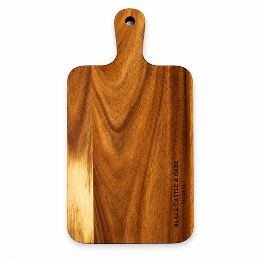 A rectangular wooden cutting board with a rounded handle and a hanging hole, shown on a clean white background. The board features a rich medium-brown wood grain with a lighter, blonde streak running vertically on the lower left, and the brand name 'BLACK CASTLE & HARE' is printed vertically on the lower right.