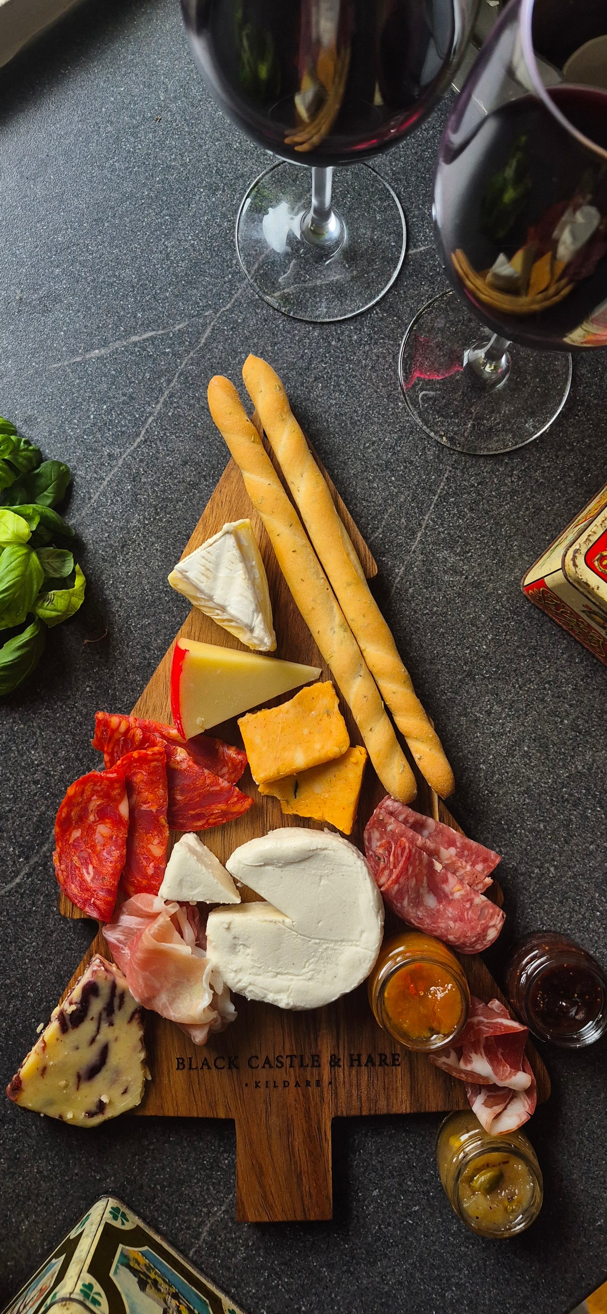 A wooden charcuterie board arranged in a tree shape with breadsticks, cheeses, cured meats, crackers, and small condiments, photographed on a dark stone surface with wine glasses partially visible in the background.