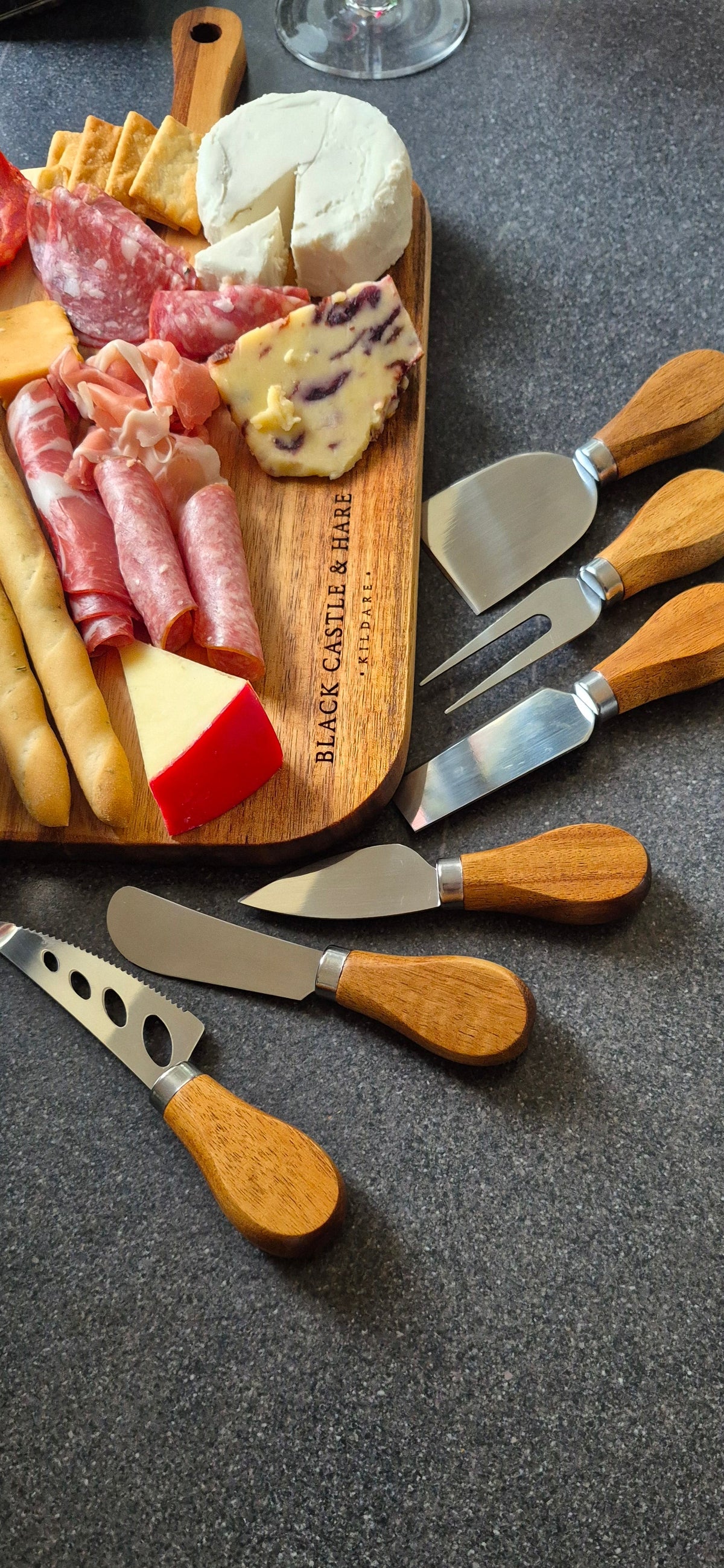 A set of wooden-handled cheese knives and spreaders arranged on a dark countertop beside a charcuterie board with cheese, salami, crackers, and bread.