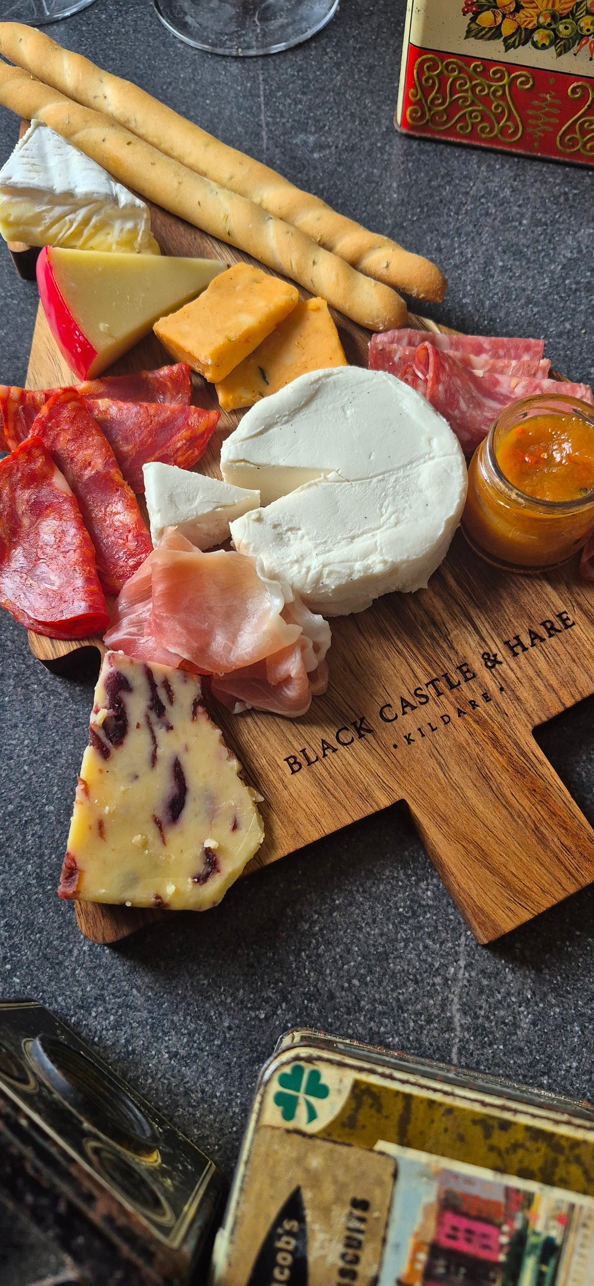 A charcuterie board styled with cheeses, cured meats, breadsticks, and a small jar of preserves on a dark textured surface. A wooden board in the center reads 'BLACK CASTLE & HARE KILDARE'.
