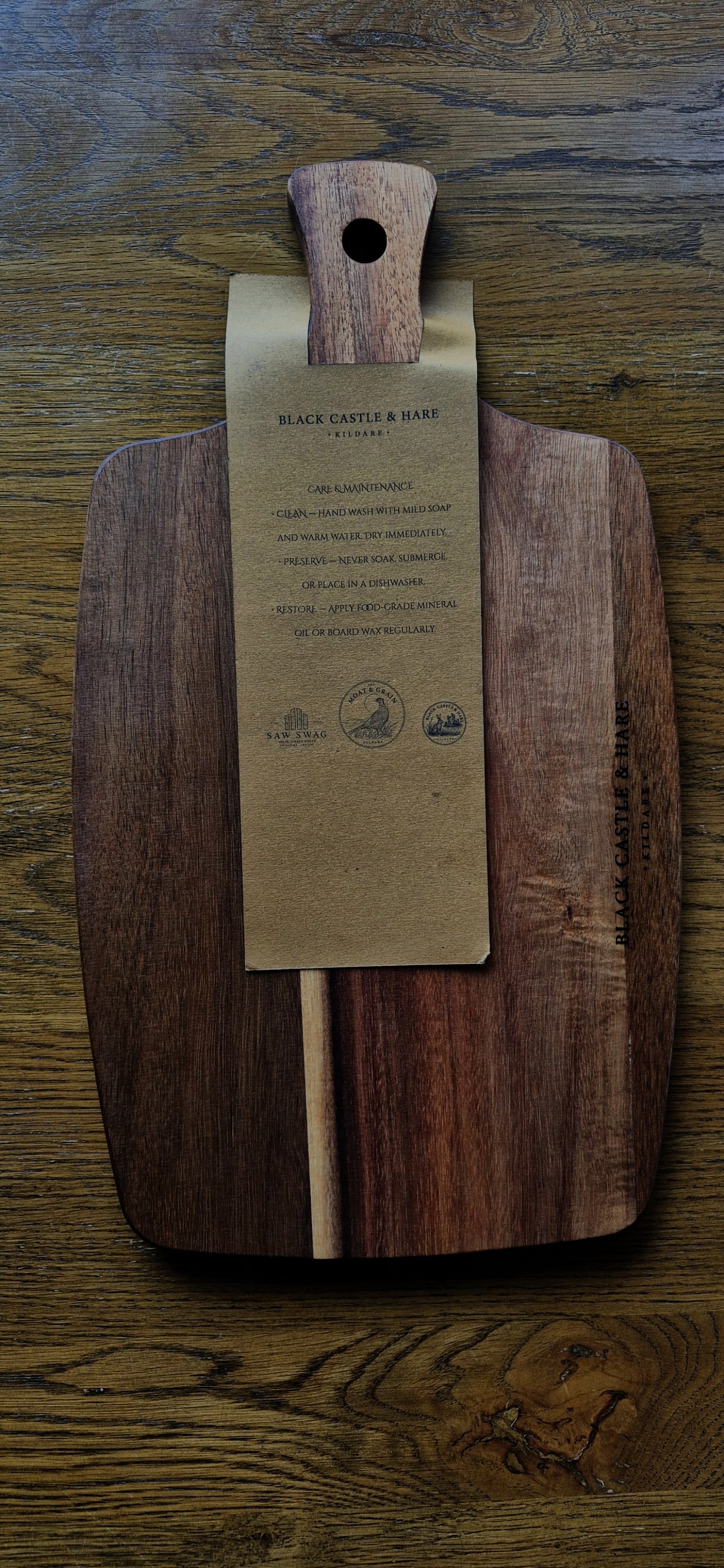 A large dark brown wooden cutting board with a natural wood handle, featuring a kraft paper tag attached to the center with board care instructions, placed on a rustic wooden table.