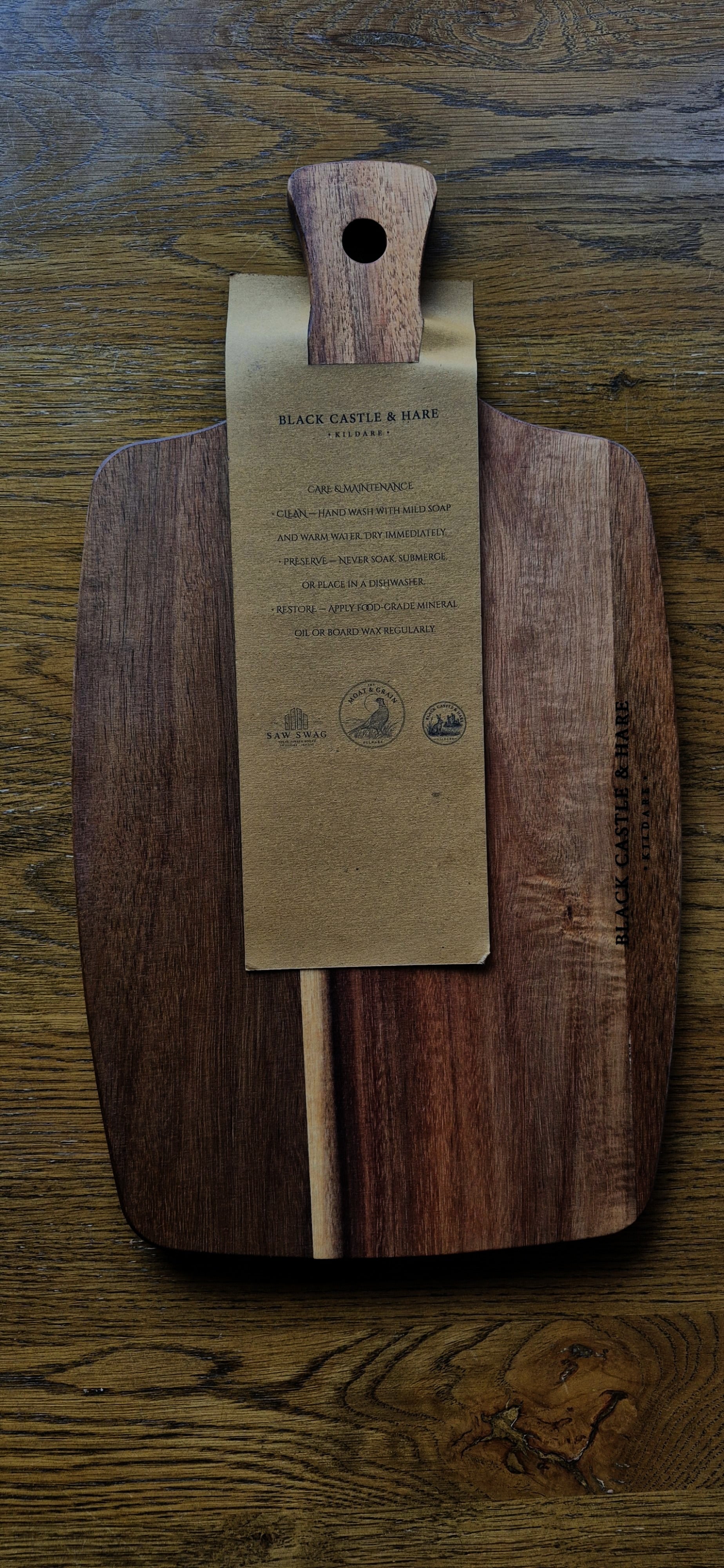 A large dark brown wooden cutting board with a natural wood handle, featuring a kraft paper tag attached to the center with board care instructions, placed on a rustic wooden table.