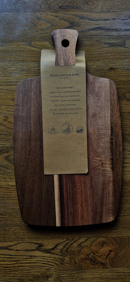 A large dark brown wooden cutting board with a natural wood handle, featuring a kraft paper tag attached to the center with board care instructions, placed on a rustic wooden table.