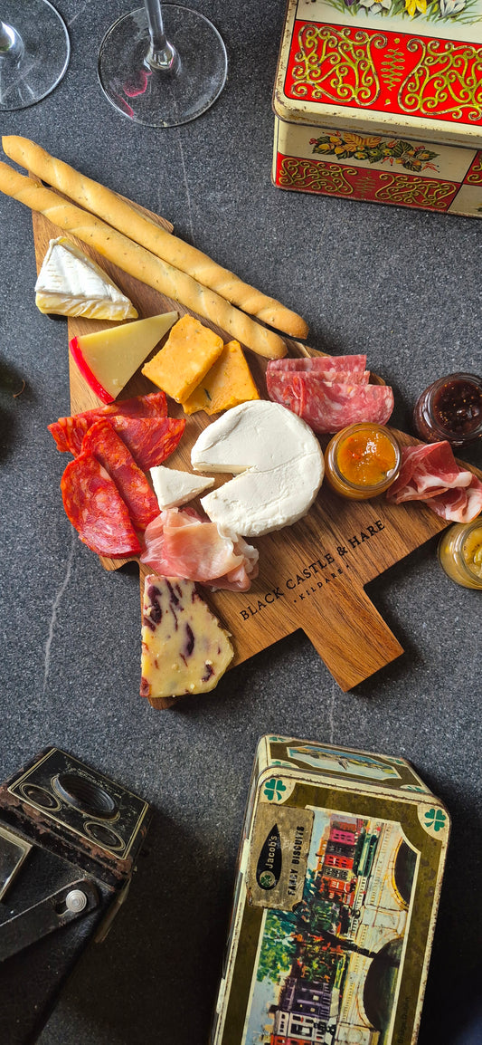 An overhead styled food spread on a dark stone surface featuring a wooden charcuterie board with cheeses, cured meats, breadsticks, crackers, and small jars, with decorative boxes and wine glasses partially visible around it.