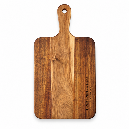 A rectangular wooden cutting board with a rounded handle and a hanging hole, featuring a vertical grain pattern and the engraved brand name 'BLACK CASTLE & HARE LONDON' on the right side, shown against a plain white background.