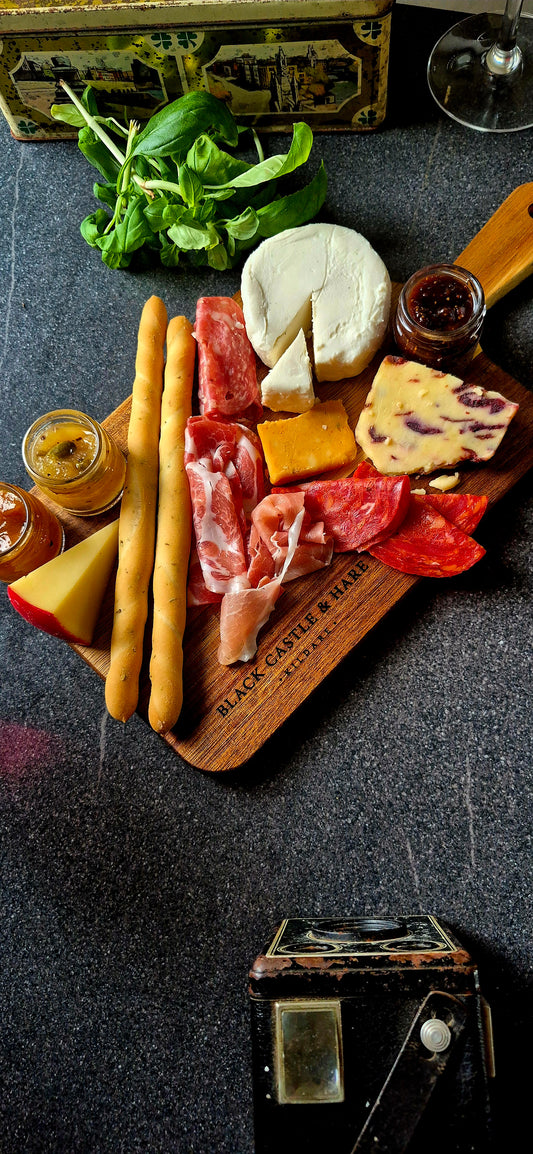 Top-down view of an artisan acacia paddle board loaded with cured meats, cheeses, and breadsticks with wine glasses in the background.