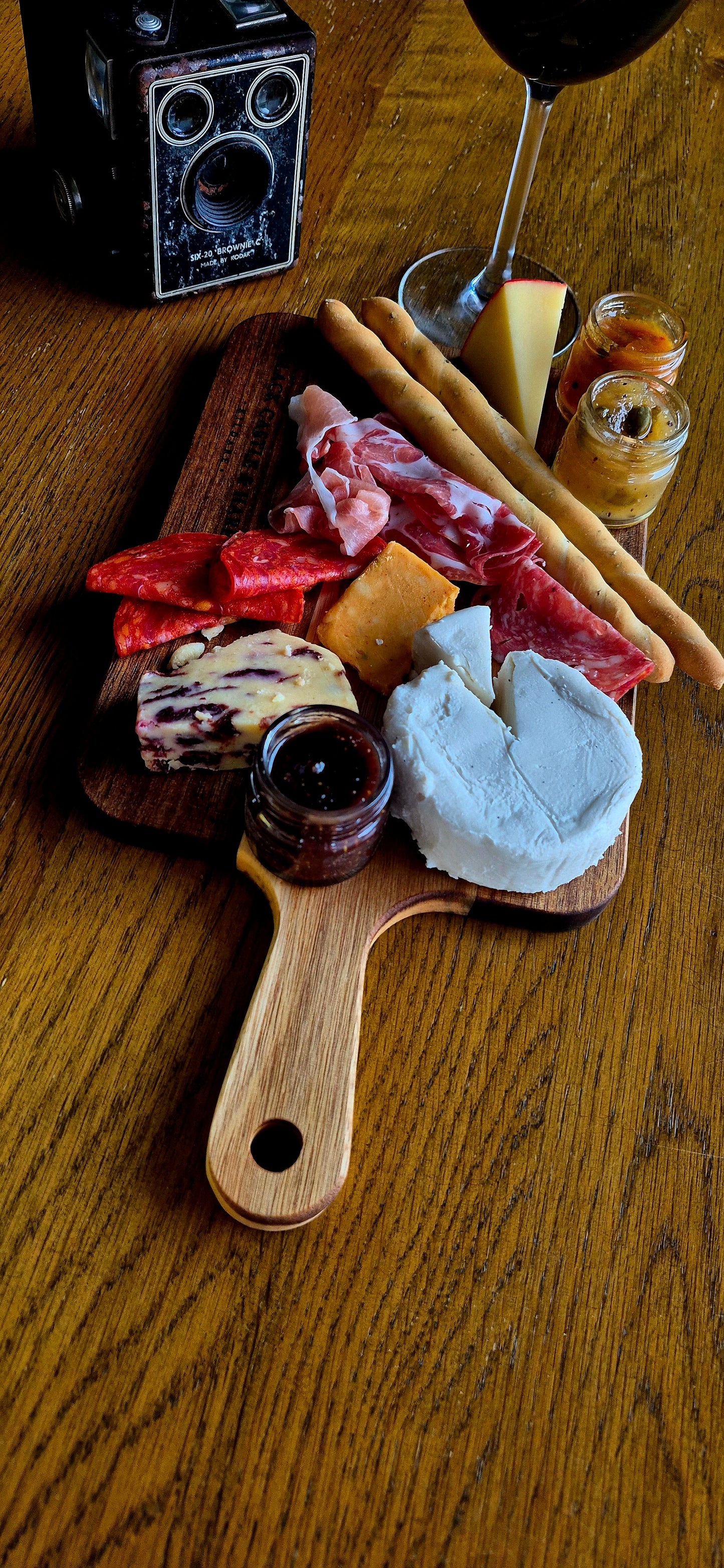 A wooden charcuterie board with an assortment of cheeses, cured meats, crackers, and small jars of spreads, arranged on a wooden table next to a wine glass and a vintage camera.