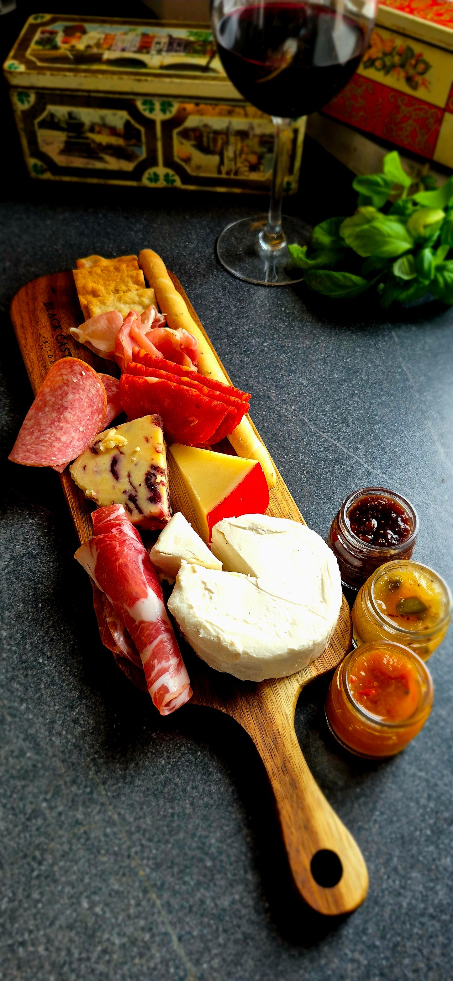 Small acacia charcuterie board styled with artisan cheese, salami, and crispy breadsticks next to a wine glass background
