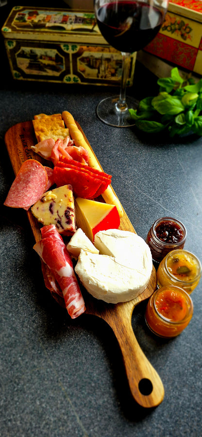 Small acacia charcuterie board styled with artisan cheese, salami, and crispy breadsticks next to a wine glass background
