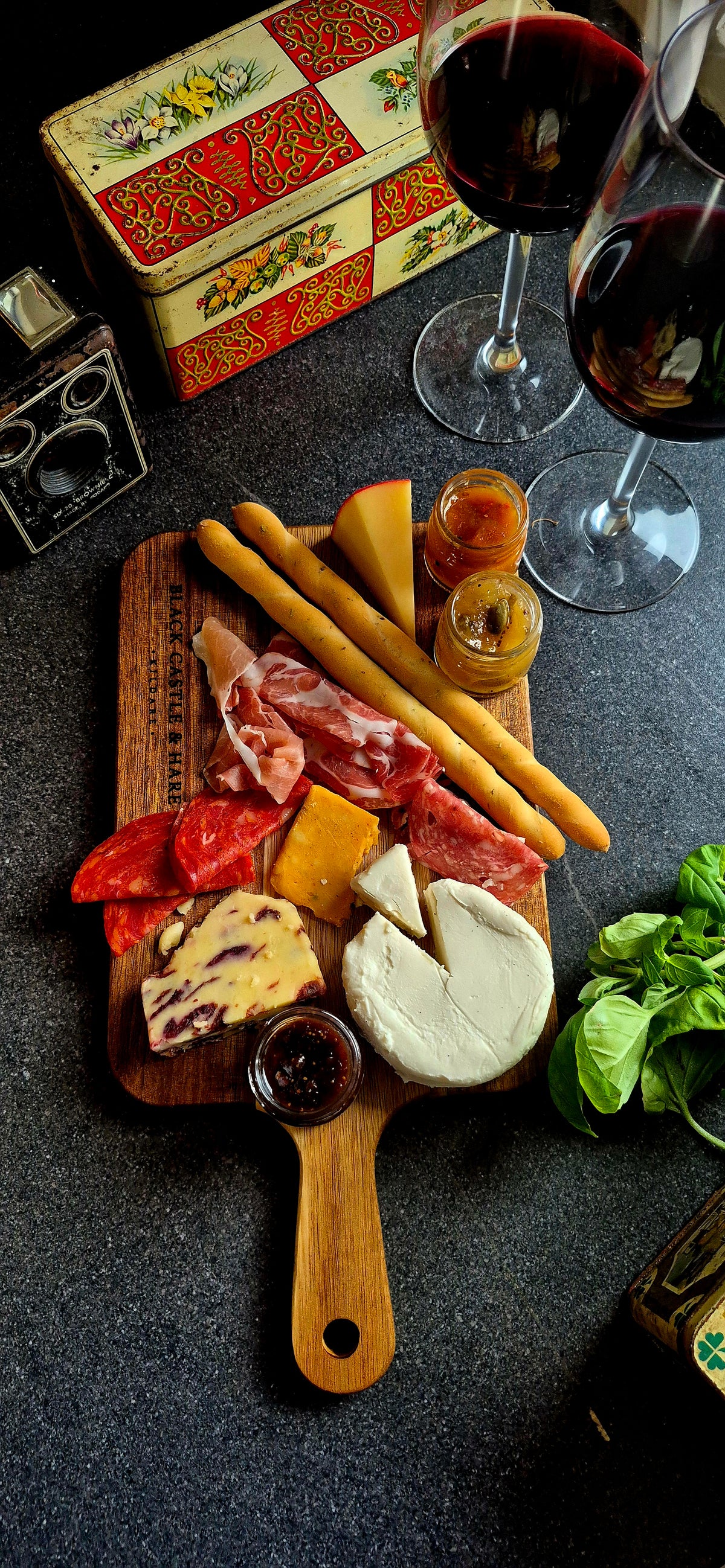 Acacia charcuterie paddle board prepared for dining, laden with cured meats, artisan cheeses, breadsticks, and wine glasses