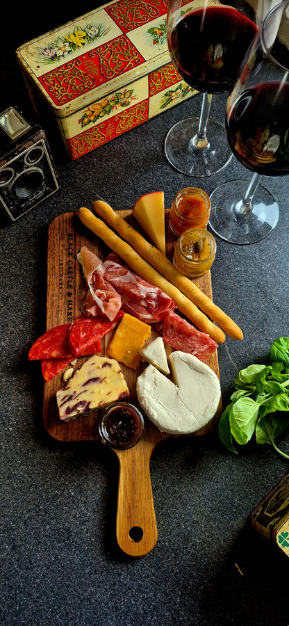 Acacia charcuterie paddle board prepared for dining, laden with cured meats, artisan cheeses, breadsticks, and wine glasses