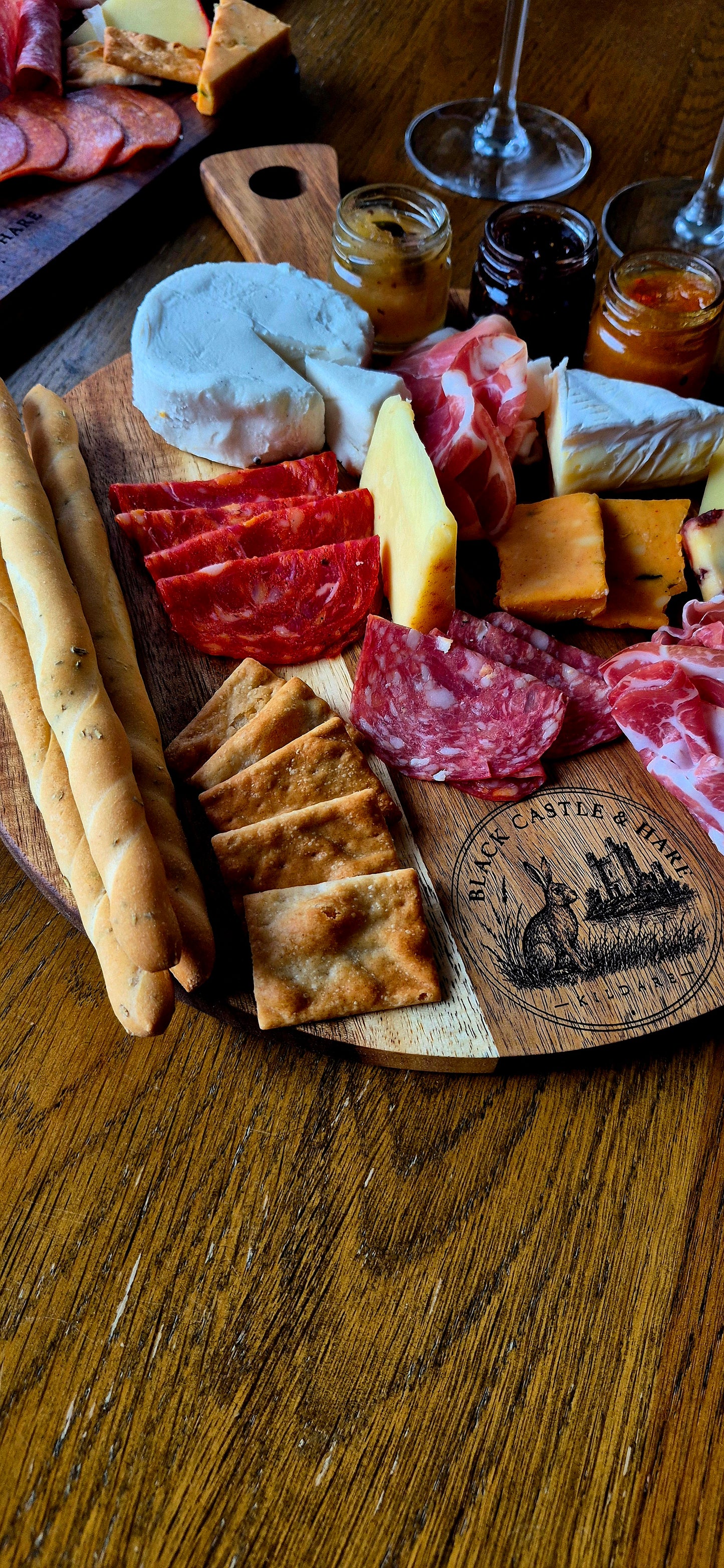 Soft-focus dining scene of double-oiled round acacia charcuterie board with cured meats, cheese, breadsticks, and wine glasses