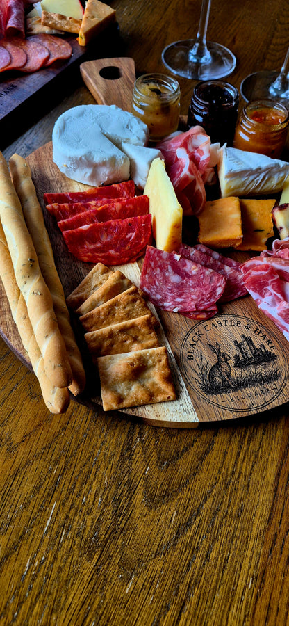 Soft-focus dining scene of double-oiled round acacia charcuterie board with cured meats, cheese, breadsticks, and wine glasses