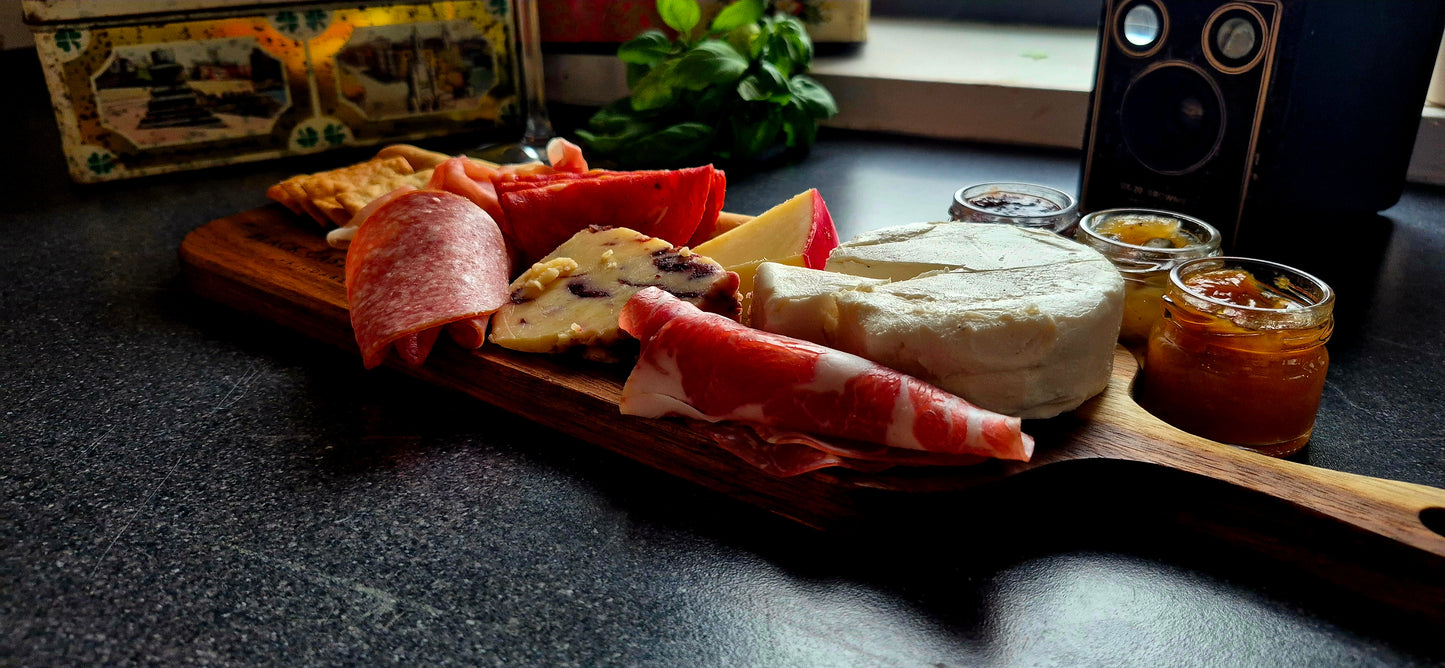 Personal-size food-safe acacia serving board featuring cured meat and cheese arrangement with wine glasses in soft focus background