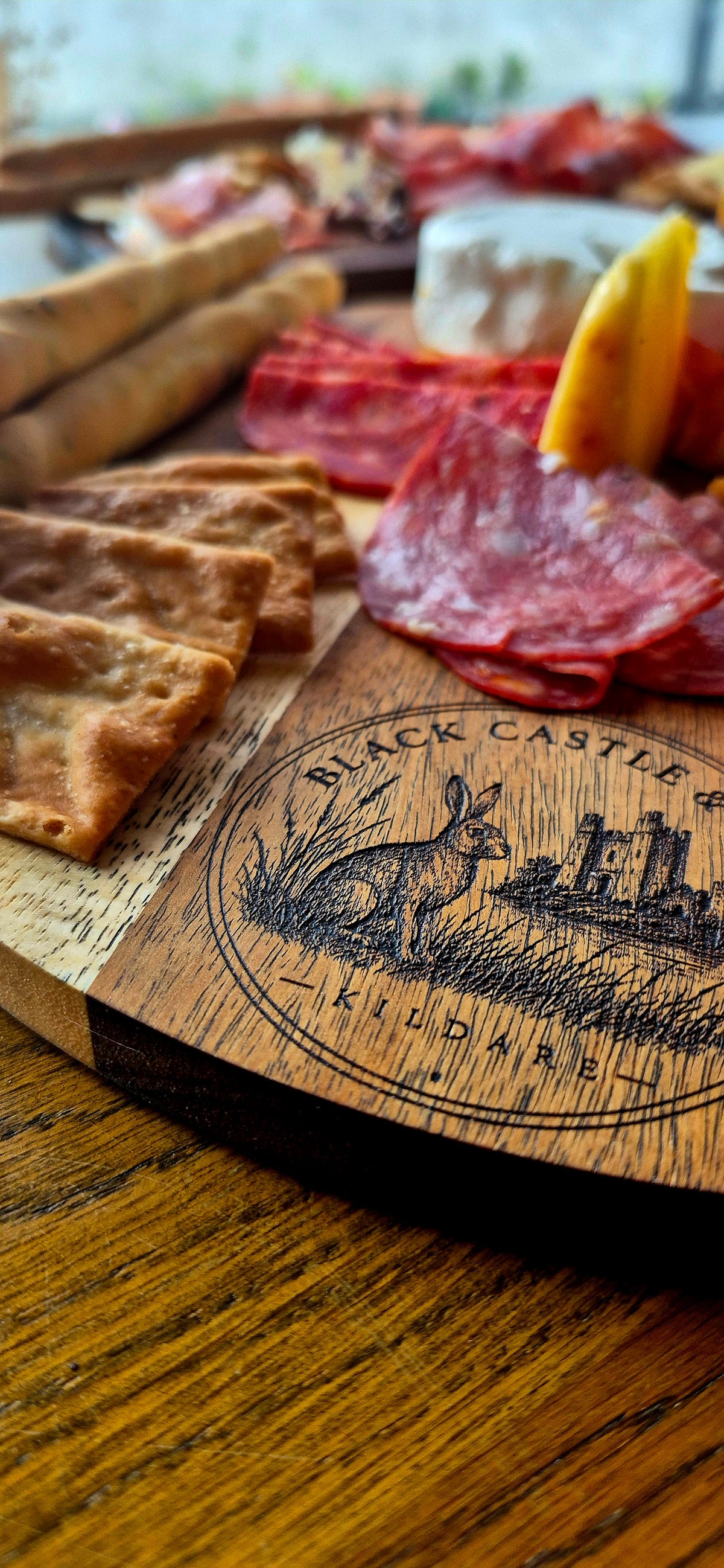 A close-up of a wooden charcuterie board engraved with 'Black Castle Killshrie' and a rabbit illustration, set on a wooden table with sliced meats, crackers, and cheese.