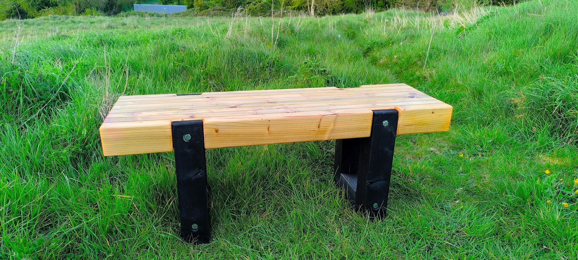 Wooden bench with black wooden legs on a grassy field Handcrafted durable wooden garden furniture built with treated and stabilized wood to last for many years.