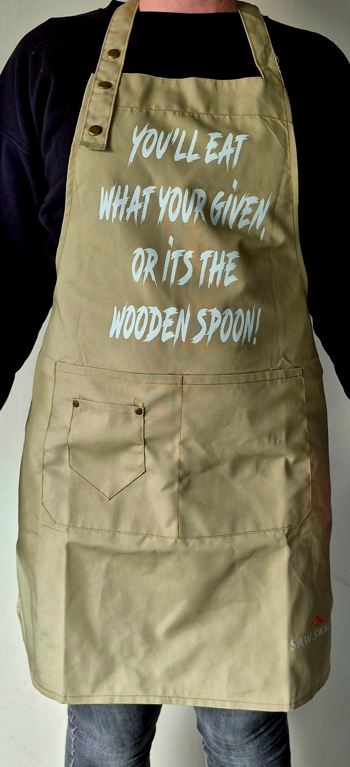 Khaki apron front view showing "Eat what you're given or it's the wooden spoon" text.