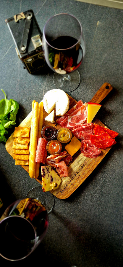Flat lay of 350x250mm acacia charcuterie board loaded with cheeses, cured meats, and breadsticks with wine glasses and an old kodak Brownie camera in background