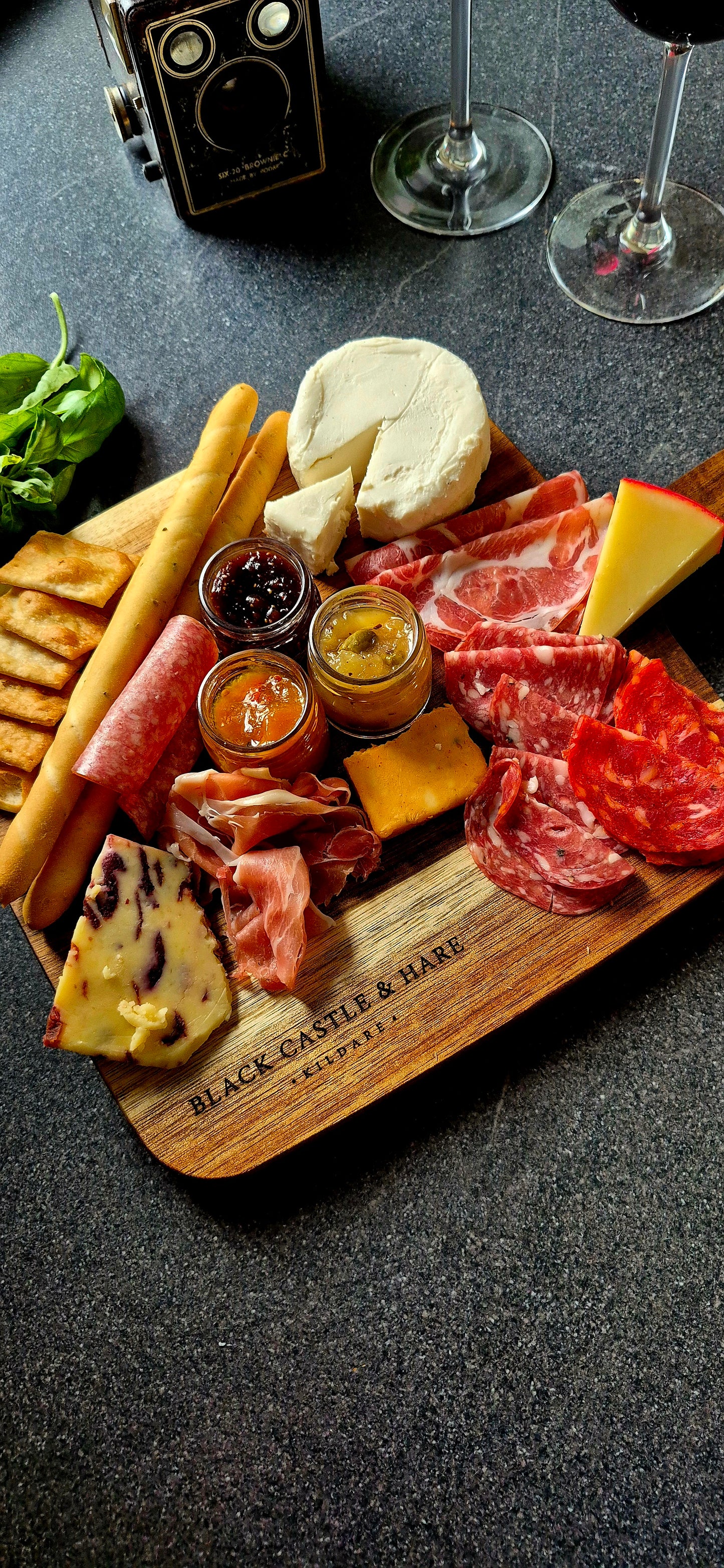 Flat lay of 350x250mm acacia charcuterie board loaded with cheeses, cured meats, and breadsticks with wine glasses in background
