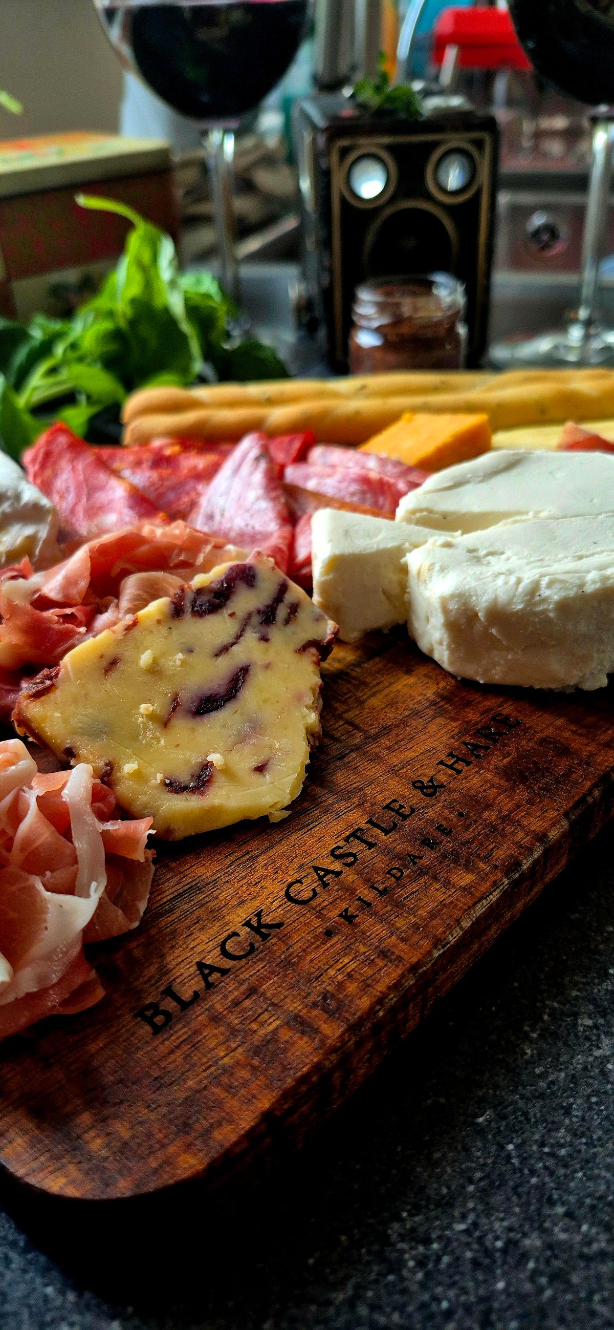 Angled view of 15mm thick acacia serving board with handle, featuring charcuterie, breadsticks, and glasses of wine in soft focus.