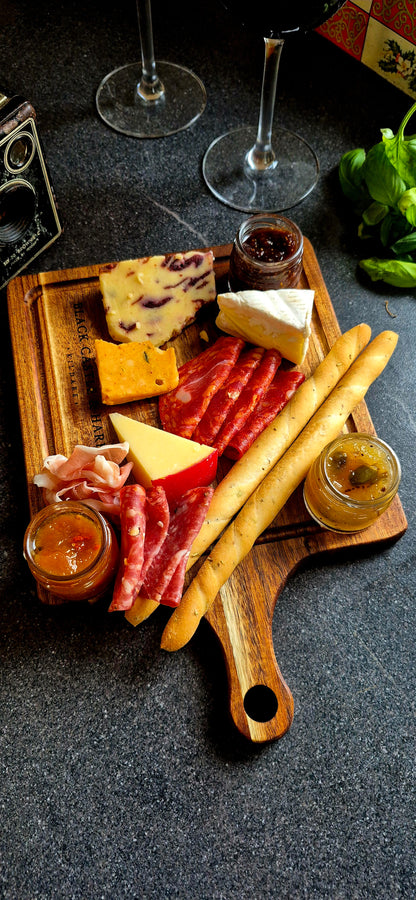 A charcuterie board styled with various cheeses, sliced cured meats, baguettes, and small jars of spreads, set on a dark textured surface with wine glasses in the background.