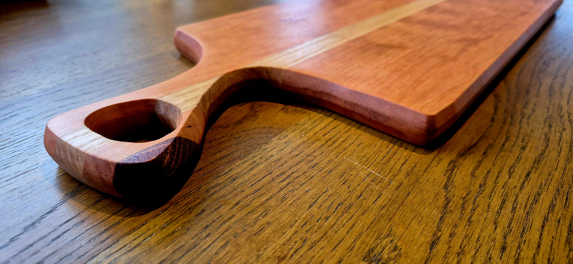 A handcrafted serving board featuring a central Oak panel wrapped in a Cherry wood frame with a dark Walnut handle accent