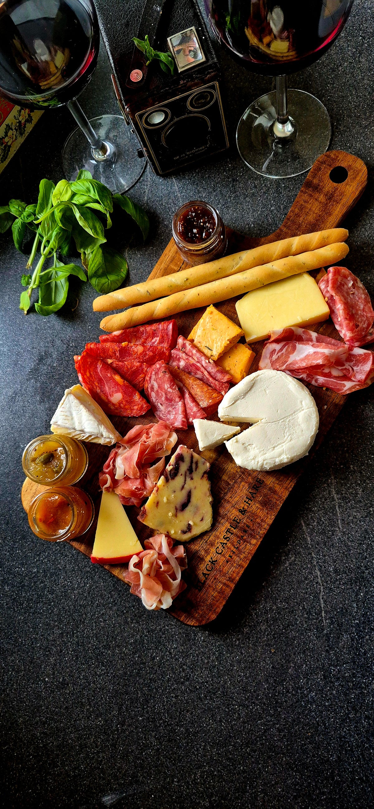 Top-down view of 395mm x 195mm acacia paddle board with handle, styled with cured meats, artisan cheese, breadsticks, and wine