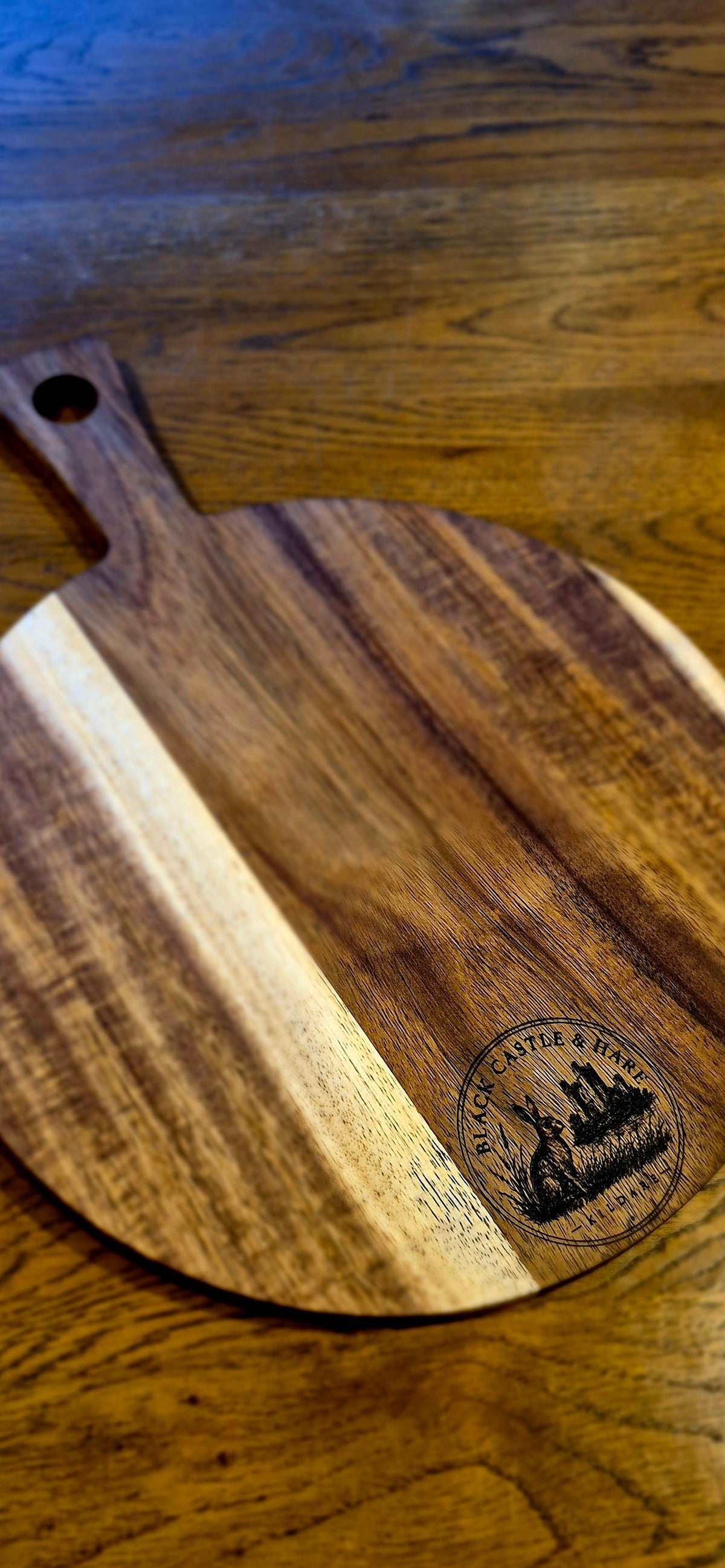 A round wooden serving board with a handle, featuring a dark and light wood grain pattern and an engraved logo, placed on a wooden tabletop.