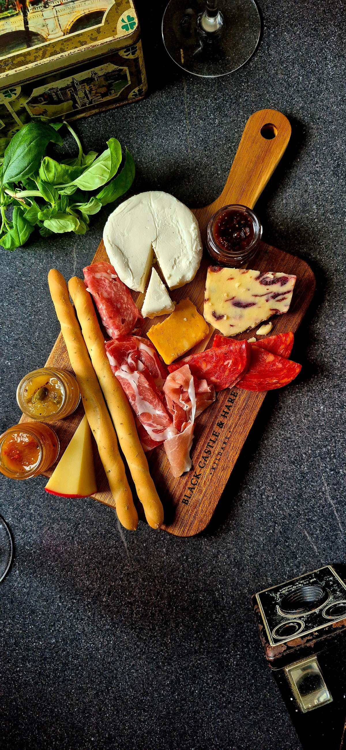 Angled view of a 15mm thick acacia serving board with handle, featuring charcuterie, crispy breadsticks, and red wine in soft focus