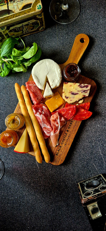 Angled view of a 15mm thick acacia serving board with handle, featuring charcuterie, crispy breadsticks, and red wine in soft focus